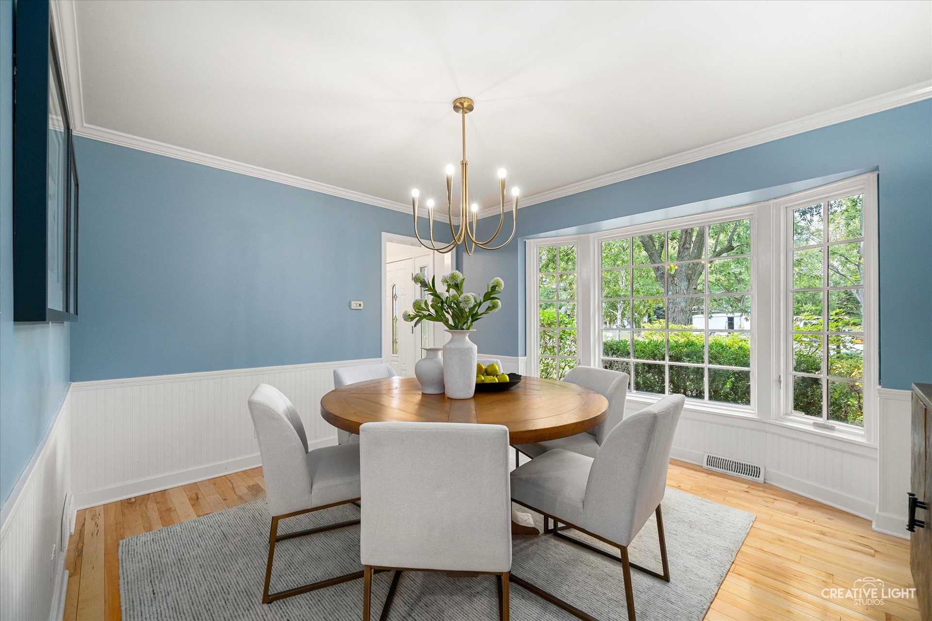 Dining room at 1101 Barberry Court Downers Grove with soft blue walls, modern chandelier, round wood table, and large bay window overlooking greenery.