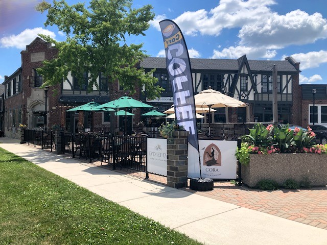 Image shows an outdoor dining area adjacent to a building with Tudor-style architecture. The building has a brick facade with large windows and decorative half-timbering. In front of the building, there is a patio area with several tables and chairs, some of which are shaded by large umbrellas. The patio is bordered by a low metal fence. A tall, vertical banner with the word "coffee" is positioned near the entrance to the patio, next to a stone pillar. There are two signs attached to the fence, one with the name "Evolet Eve" and the other with the name "Cora" accompanied by a logo. A grassy area runs alongside the sidewalk leading to the patio. A large tree provides additional shade to the area.