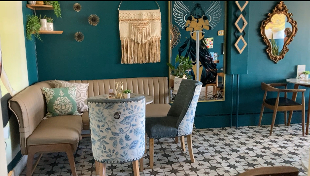 A seating area in the salon of Evolet Eve and Cora Coffee. Thee is a round table with a floral-patterned chair on the left and a plain chair on the right. The floor has a patterned tile design. The wall int he background is adorned with a macrame wall hanging and a large decorative piece featuring an eagle with outstretched wings.