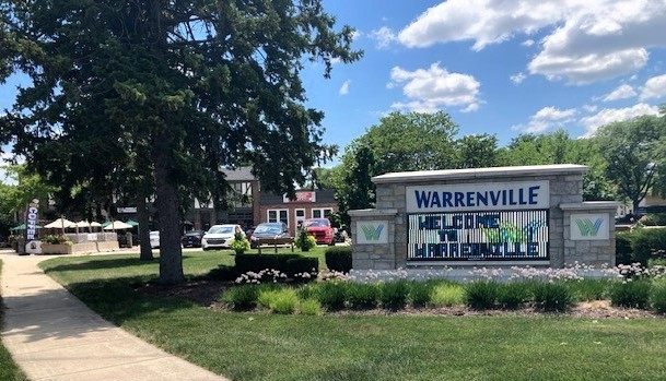 The image shows a sign for Warrenville, which is prominently displayed in the center-right of the image. The sign is constructed from stone and features the text "Warrenville" at the top, with "Welcome to" and "Warrenville" below it. The sign is surrounded by a well-maintained garden with various plants and flower in front of it.
